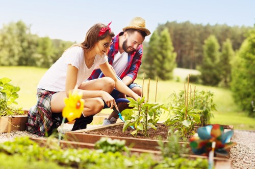 A young girl and a man are working together in a well-maintained garden, planting or tending to a vegetable bed with small green plants. The girl has long brown hair and is wearing a white t-shirt, plaid shorts, and a pink headband with a decorative bow, while the man is wearing a straw hat, a checkered shirt, and jeans. They are kneeling on the gravel pathway that runs alongside the raised wooden garden bed, which is filled with rich soil and young plants. In the foreground, there are additional garden beds with leafy plants, and a small yellow decorative garden ornament is visible nearby. The background features a lush, green lawn, mature trees, and a clear blue sky, suggesting a sunny day in a suburban or rural setting. This scene highlights outdoor gardening activities typical of a private garden in Hackbridge and showcases the natural tones, textured soil, and vibrant green foliage common in local landscaping efforts and garden maintenance services provided by companies like Hedge Trimming Hackbridge.