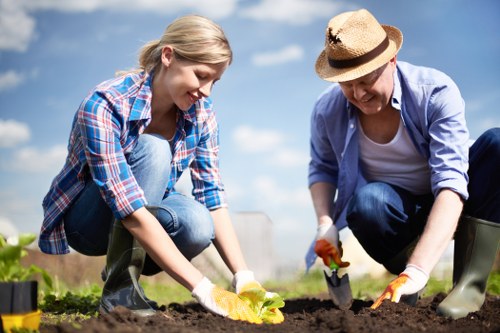A young woman and an older woman are working together outdoors in a well-maintained garden, planting new plants into the soil. The younger woman, wearing a blue checkered shirt and jeans, is crouched down, carefully placing a young plant into the ground, while the older woman, dressed in a light blue jacket, white shirt, and a wide-brimmed straw hat, is also bent over, using gardening gloves and a small trowel to assist with planting. The garden features a neatly dug flower bed with freshly tilled dark soil, bordered by a paved pathway, and visible green leafy plants nearby. The background shows a bright, partly cloudy sky, suggesting daytime with natural sunlight illuminating the scene, typical of gardening activities in Hackbridge, south London. This outdoor setting highlights elements common in domestic gardens, including cultivated flower beds, healthy soil, and the collaborative effort involved in garden maintenance and planting, exemplifying services offered by Hedge Trimming Hackbridge in gardening and outdoor landscaping.