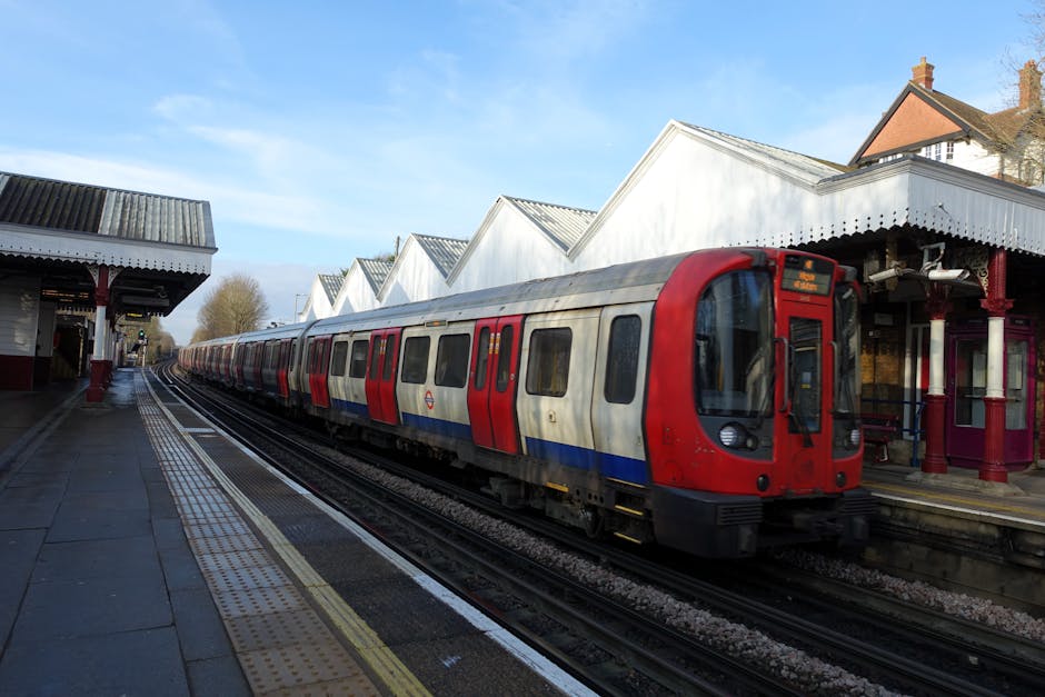 A modern train with a red front and silver body featuring red and blue stripes runs along railway tracks at a rural station platform. The platform is paved with dark tiles and has tactile paving strip